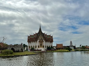 A traditional Thai temple with ornate architecture is situated by a calm body of water. The temple is surrounded by lush greenery and a stone bridge, while a clear sky with scattered clouds forms the backdrop.