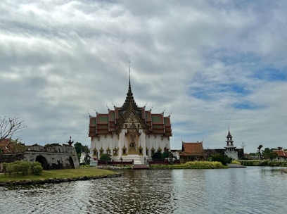 A traditional Thai temple with ornate architecture is situated by a calm body of water. The temple is surrounded by lush greenery and a stone bridge, while a clear sky with scattered clouds forms the backdrop.