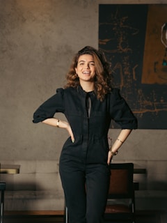 A woman with curly hair is standing confidently, wearing a black jumpsuit with her hands on her hips. She has a cheerful expression on her face and is positioned in an indoor setting with a couch and a piece of abstract art hanging on the wall behind her.