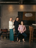 Three people are posing together in a stylish and casually arranged space. The setting includes a textured wall and a piece of modern art in the background. They are standing near a few chairs and wooden tables, creating a relaxed and friendly atmosphere.