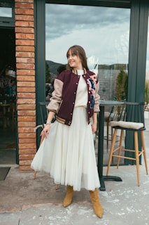 A person stands outdoors wearing a maroon and beige varsity jacket over a white dress, paired with tan suede boots. The setting includes a brick wall and a glass door with a view of trees and a cloudy sky.