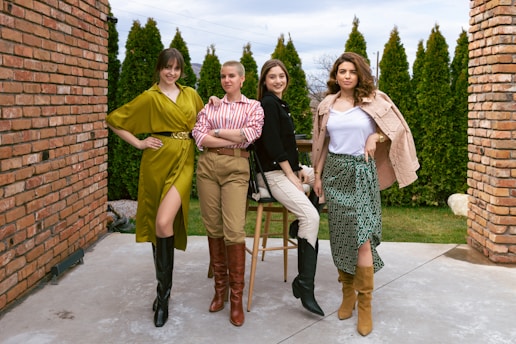 Four women are posing in stylish outfits, standing outdoors between two brick walls with a backdrop of tall green shrubs and a cloudy sky. They are standing on a paved surface, exuding confidence and camaraderie.