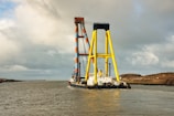A large maritime crane vessel navigates through calm waters. The structure features colorful yellow and orange latticework visible against a cloudy sky. Some land and a few birds are discernible in the distance.