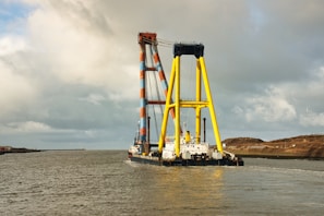 A large maritime crane vessel navigates through calm waters. The structure features colorful yellow and orange latticework visible against a cloudy sky. Some land and a few birds are discernible in the distance.