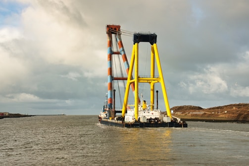 A large maritime crane vessel navigates through calm waters. The structure features colorful yellow and orange latticework visible against a cloudy sky. Some land and a few birds are discernible in the distance.
