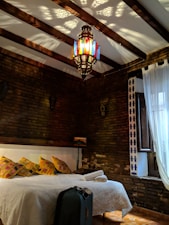 A cozy bedroom with a rustic brick wall, adorned with a colorful hanging lantern casting intricate patterns on the white ceiling. A bed is neatly made with white linens, yellow accent pillows, and a rolled-up towel. A black suitcase is placed at the foot of the bed. A window with sheer white curtains allows light to filter into the room, providing a warm and inviting ambiance. A small wooden shelf above the bed holds a decorative lamp.