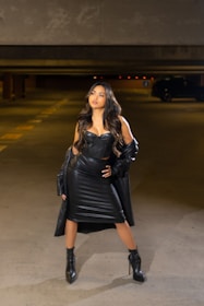 A woman stands confidently in a dimly lit parking garage, wearing an all-black leather outfit that includes a corset top, skirt, and high-heeled boots. Her long hair cascades down her shoulders, and she poses with one hand on her hip and the other holding her coat. The concrete walls and floor create a contrasting backdrop, emphasizing the sleekness and style of her attire.