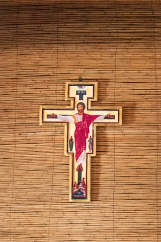 A crucifix with an image of Jesus Christ in red and white robes, surrounded by illustrations of several other figures. The background consists of horizontal wooden slats, likely forming a wall.