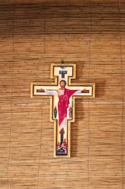 A crucifix with an image of Jesus Christ in red and white robes, surrounded by illustrations of several other figures. The background consists of horizontal wooden slats, likely forming a wall.