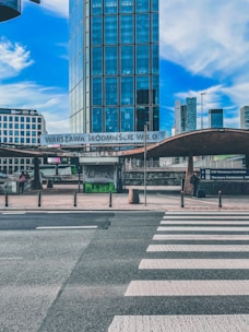 A modern cityscape featuring a tall glass skyscraper in the background with a clear blue sky. In the foreground, there is a crosswalk leading to a train station entrance labeled 'Warszawa Śródmieście W.K.D.' with several signs and advertisements. A few people are visible walking near the entrance.