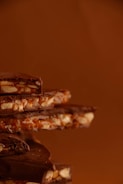 A stack of chocolate pieces mixed with nuts and caramel, against a warm brown background. The chocolate appears rich and glossy, with textures that suggest a crunchy and creamy composition.