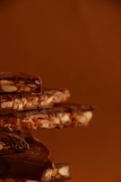 A stack of chocolate pieces mixed with nuts and caramel, against a warm brown background. The chocolate appears rich and glossy, with textures that suggest a crunchy and creamy composition.