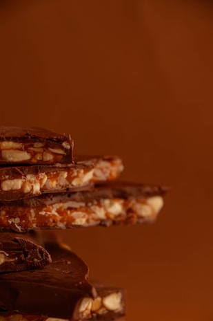 A stack of chocolate pieces mixed with nuts and caramel, against a warm brown background. The chocolate appears rich and glossy, with textures that suggest a crunchy and creamy composition.