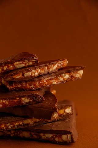A stack of chocolate bars with visible chunks of nuts embedded in them. The bars have a rich brown color and smooth surface, with nuts offering a crunchy texture. The background is a warm earthy tone, complementing the chocolate.