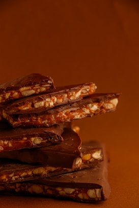 A stack of chocolate bars with visible chunks of nuts embedded in them. The bars have a rich brown color and smooth surface, with nuts offering a crunchy texture. The background is a warm earthy tone, complementing the chocolate.
