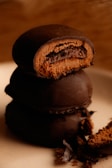A delectable stack of round chocolate-covered cookies with one partially bitten, revealing a rich inner layer of chocolate filling. Crumbs are scattered around the base, adding a rustic feel.