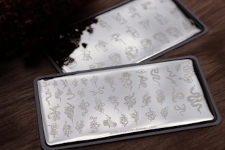 Two rectangular metallic plates engraved with intricate designs, including various animals, flowers, and abstract shapes, are placed on a wooden surface. The designs are finely detailed and reflective, catching light at certain angles.