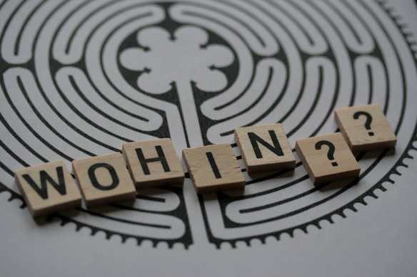 Wooden tiles with letters spelling 'WOHIN?' are placed on a printed maze-like design, creating a sense of direction and inquiry.