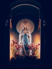 A statue of a woman wearing a crown and a long robe, standing with hands in prayer. The statue is surrounded by vibrant pink flowers and is illuminated by subtle lighting that creates a serene atmosphere. A halo effect is present behind the statue, enhancing its divine appearance.
