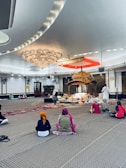 Several people sit on a carpeted floor in a spacious room with a large ornate chandelier and a decorated platform at the center. The individuals wear head coverings and face toward the platform, which is adorned with flowers and an intricately designed canopy. The setting appears peaceful and reverent.