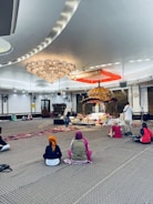 Several people sit on a carpeted floor in a spacious room with a large ornate chandelier and a decorated platform at the center. The individuals wear head coverings and face toward the platform, which is adorned with flowers and an intricately designed canopy. The setting appears peaceful and reverent.