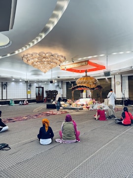 Several people sit on a carpeted floor in a spacious room with a large ornate chandelier and a decorated platform at the center. The individuals wear head coverings and face toward the platform, which is adorned with flowers and an intricately designed canopy. The setting appears peaceful and reverent.