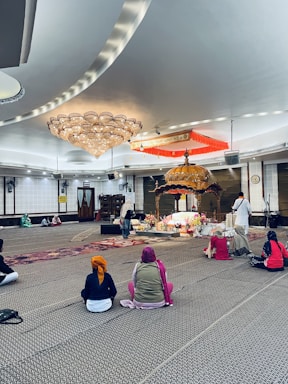 Several people sit on a carpeted floor in a spacious room with a large ornate chandelier and a decorated platform at the center. The individuals wear head coverings and face toward the platform, which is adorned with flowers and an intricately designed canopy. The setting appears peaceful and reverent.