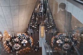 A large, modern indoor space with a high ceiling and concrete walls. There are numerous tables arranged in long rows with people seated around them, suggesting the setting might be a restaurant or a large café. On the right side, a reflection of a person appears on the glass barrier. Below, there are multiple vendor booths and display tables, possibly indicating a market or exhibition. The area is bustling with activity, and there are Christmas trees or decorative wooden structures visible.