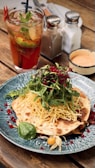 A well-presented dish on a blue patterned plate, featuring a flatbread topped with shredded cheese, fresh greens, and pomegranate seeds. Next to the plate, a glass of iced tea with mint and lime, along with salt and pepper shakers, are arranged on a rustic wooden table. A small dish of sauce accompanies the ensemble.