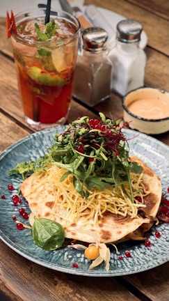 A well-presented dish on a blue patterned plate, featuring a flatbread topped with shredded cheese, fresh greens, and pomegranate seeds. Next to the plate, a glass of iced tea with mint and lime, along with salt and pepper shakers, are arranged on a rustic wooden table. A small dish of sauce accompanies the ensemble.