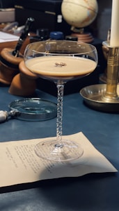A coupe glass filled with an espresso martini sits on a piece of paper. The drink is garnished with a few coffee beans. Surrounding the glass are various items on a dark table, including a magnifying glass, some wooden objects, a globe, a candle, and a pen. The lighting casts soft shadows, emphasizing the texture of the glass and the creamy surface of the cocktail.