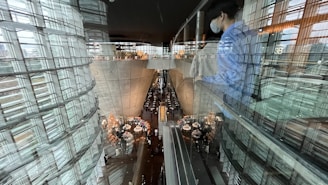 A modern, futuristic interior with large curved glass and concrete structures is visible. People are gathered around tables in a spacious area below, suggesting a restaurant setting. Reflections on the glass and a person wearing a blue shirt and face mask can also be seen.