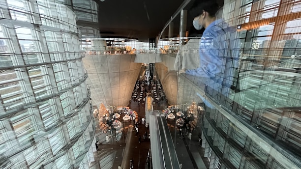 A modern, futuristic interior with large curved glass and concrete structures is visible. People are gathered around tables in a spacious area below, suggesting a restaurant setting. Reflections on the glass and a person wearing a blue shirt and face mask can also be seen.