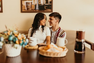 A couple is sitting closely on a sofa in a cozy setting. On the table in front of them, there are cups of coffee, a vase with colorful flowers, and a basket with pastries. The background includes framed pictures on the wall.
