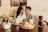 A couple is sitting closely on a sofa in a cozy setting. On the table in front of them, there are cups of coffee, a vase with colorful flowers, and a basket with pastries. The background includes framed pictures on the wall.