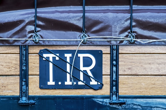 A close-up view of a wooden and metal structure with a sign that has the letters 'T.I.R' on it. The structure is part of a covered vehicle with a tarpaulin attached using metal hooks and ropes.