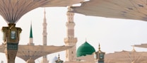 Several large architectural umbrellas are open against a backdrop of a mosque with ornate minarets and a green dome. The structures are intricately designed and arranged to provide shade.