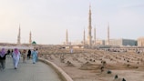 A serene landscape captures several towering minarets and a large expanse of a cemetery in the foreground. People, some dressed in traditional attire, walk along a paved pathway bordering the cemetery. The minarets belong to a grand mosque visible in the distance, with the smooth transitions of a light blue sky overhead.
