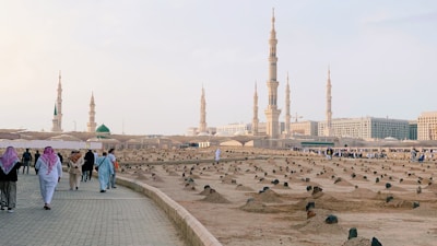 A serene landscape captures several towering minarets and a large expanse of a cemetery in the foreground. People, some dressed in traditional attire, walk along a paved pathway bordering the cemetery. The minarets belong to a grand mosque visible in the distance, with the smooth transitions of a light blue sky overhead.