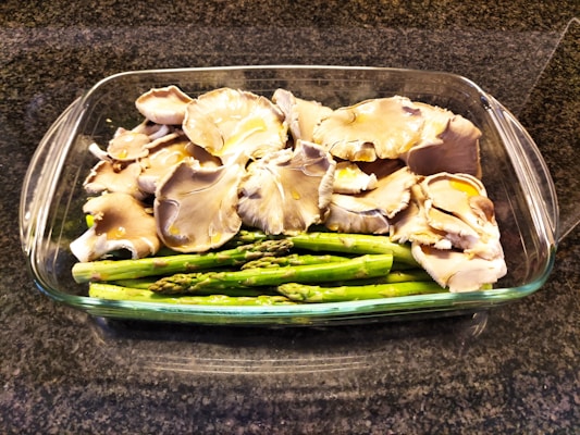 A glass baking dish contains a neatly arranged bed of fresh asparagus topped with a layer of raw oyster mushrooms. The dish is placed on a dark granite countertop.