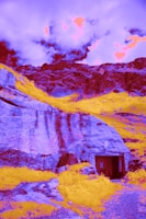 Surreal landscape with vibrant colors featuring rugged mountains and a cave-like entrance. The sky is dramatically lit with intense, psychedelic hues.