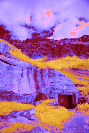 Surreal landscape with vibrant colors featuring rugged mountains and a cave-like entrance. The sky is dramatically lit with intense, psychedelic hues.