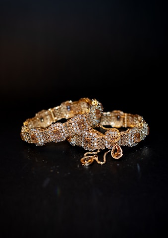 A pair of intricately designed golden bracelets adorned with small sparkling gems. Each bracelet features a symmetrical pattern with alternating diamond-shaped sections and embedded gemstones, connected by a decorative clasp and a dangling teardrop-shaped charm.