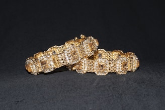 Two intricately crafted gold bracelets with embedded sparkling gemstones are displayed against a dark background. The bracelets are ornate, with a pattern of square shapes and shimmering surfaces, suggesting luxury and elegance.