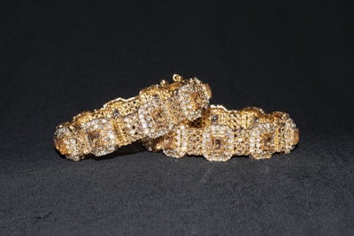 Two intricately crafted gold bracelets with embedded sparkling gemstones are displayed against a dark background. The bracelets are ornate, with a pattern of square shapes and shimmering surfaces, suggesting luxury and elegance.
