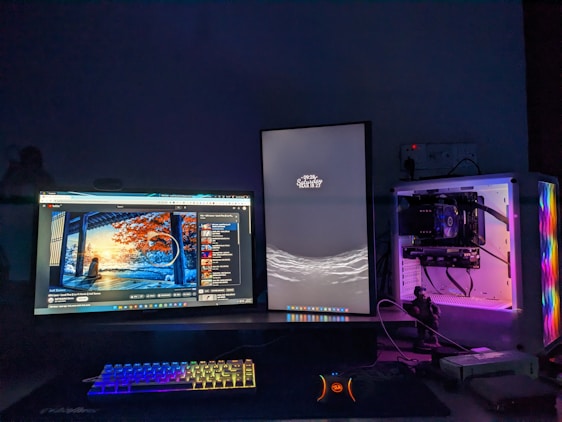 A computer setup featuring two monitors is displayed. The left monitor shows a colorful digital artwork of a serene landscape scene with trees and a sunrise or sunset. The right monitor is vertically oriented with a minimalist design showing the date and time. A compact keyboard with rainbow LED backlighting is placed beneath the monitors. A white computer case with transparent panels is visible on the right, illuminated by internal RGB lighting.