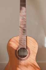 A classical acoustic guitar displaying its wooden body and intricate rosette around the sound hole. Sunlight casts soft shadows on the guitar, highlighting its smooth texture and craftsmanship. The strings and fretboard are clearly visible, emphasizing the instrument's elegant design.