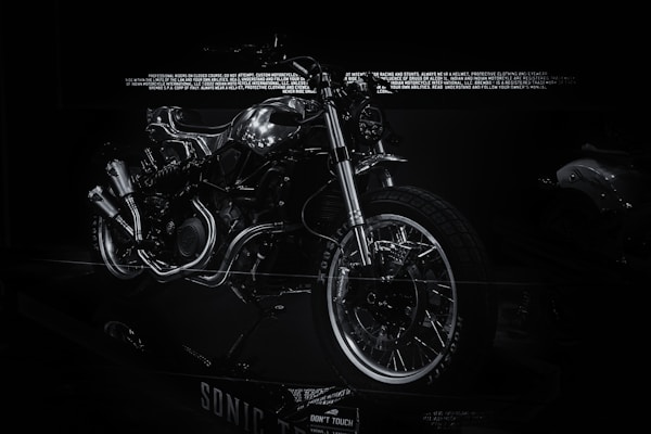 A futuristic and sleek motorcycle is prominently displayed with a predominantly metallic design, highlighted by reflective chrome parts. The bike features exposed mechanical components, giving it an industrial aesthetic. The surrounding environment is dark, making the motorcycle the focal point as light reflects off its metallic surfaces.
