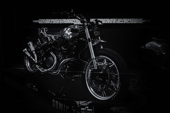 A futuristic and sleek motorcycle is prominently displayed with a predominantly metallic design, highlighted by reflective chrome parts. The bike features exposed mechanical components, giving it an industrial aesthetic. The surrounding environment is dark, making the motorcycle the focal point as light reflects off its metallic surfaces.