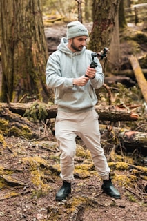 A person wearing a gray hoodie and beanie stands in a forested area. They are holding a gimbal, focusing on capturing footage or taking photos. The background features thick tree trunks and a forest floor covered with moss and leaves.
